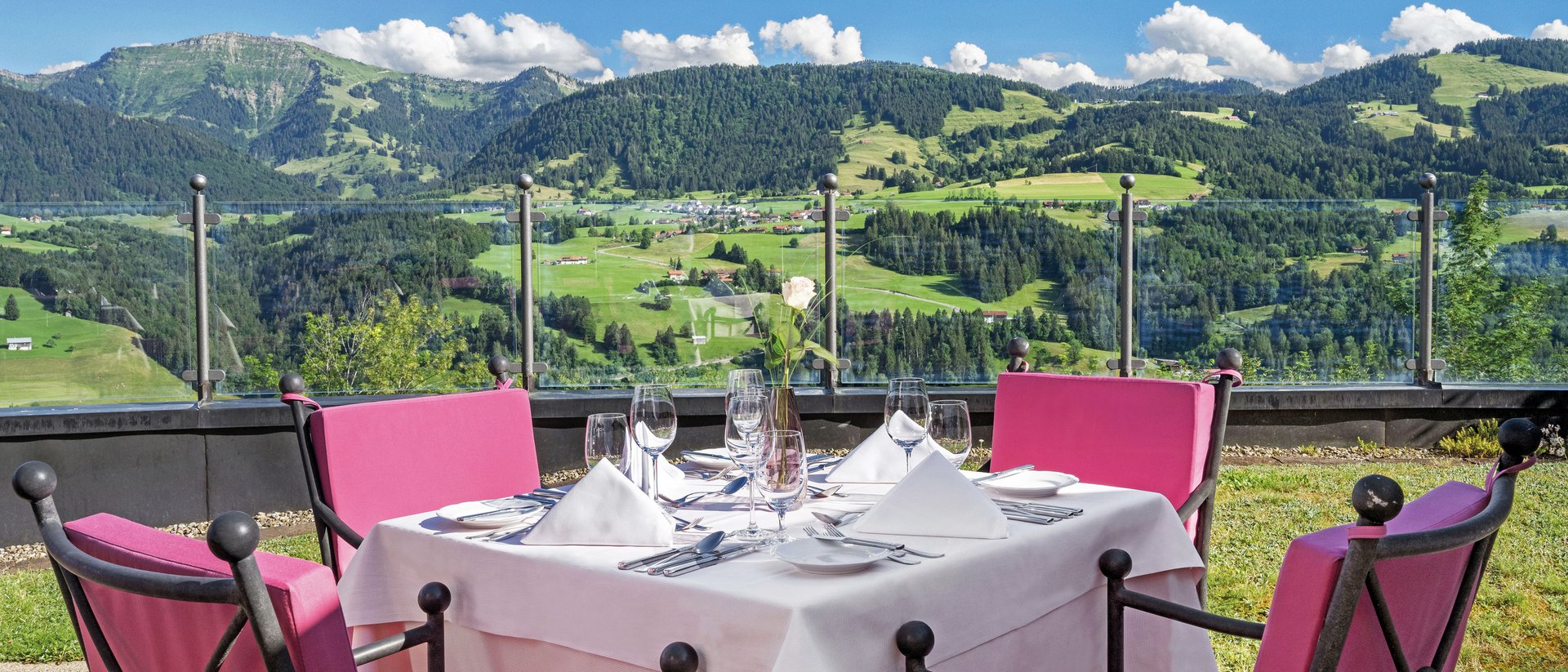 Oberstaufen: Restaurant mit 5-Sterne-Küche Outdoor-Tisch mit weißen Tüchern und pinken Stühlen vor Bergkulisse bei Sonnenschein