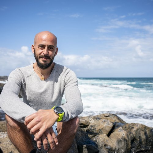 Wissen, das inspiriert: Unsere Vortragsreihe greift Themen auf, die das Leben bereichern. Mann mit Bart und grauem Pullover sitzt auf Felsen am Meer bei sonnigem Wetter