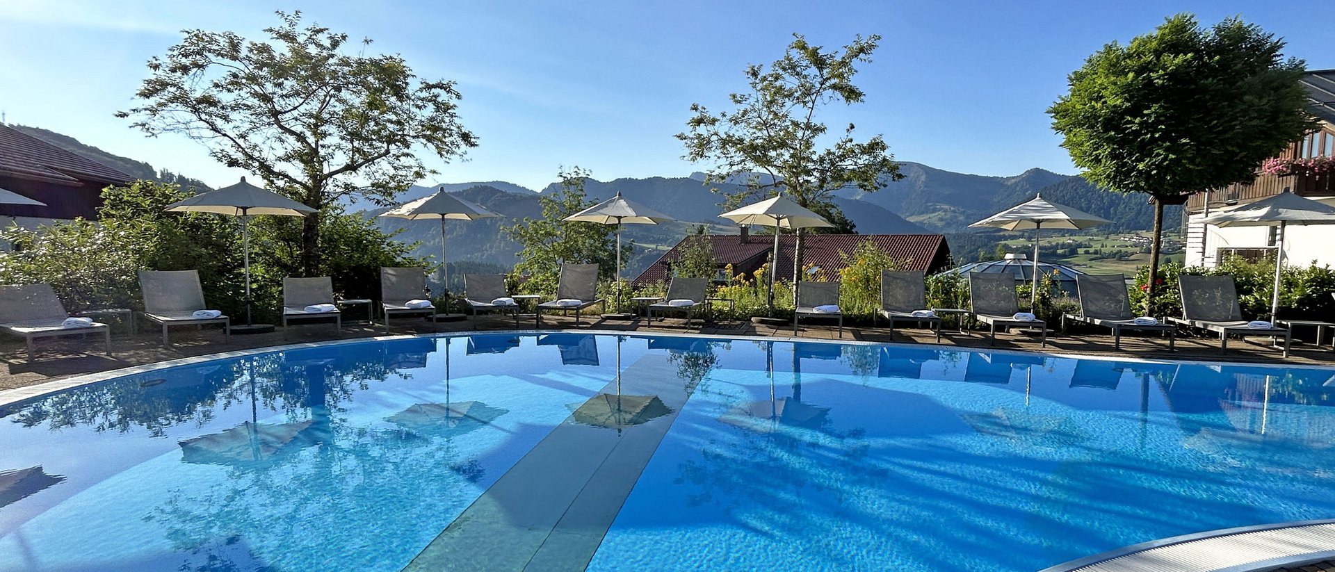 Hotel mit Pool im Allgäu: Allgäu Sonne Leerer Pool mit Sonnenschirmen und Liegestühlen vor Berglandschaft