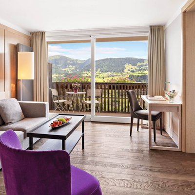 Original Schrothkur in Oberstaufen im Hotel Allgäu Sonne Helles Wohnzimmer mit Sofa, lila Sessel, Schreibtisch und Balkon mit Bergblick